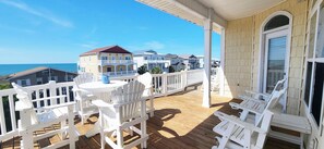 Eine Holzterrasse mit weißen Adirondack-Stühlen und einem Tisch, mit Blick auf ein Wohngebiet mit Häusern und dem Meer.