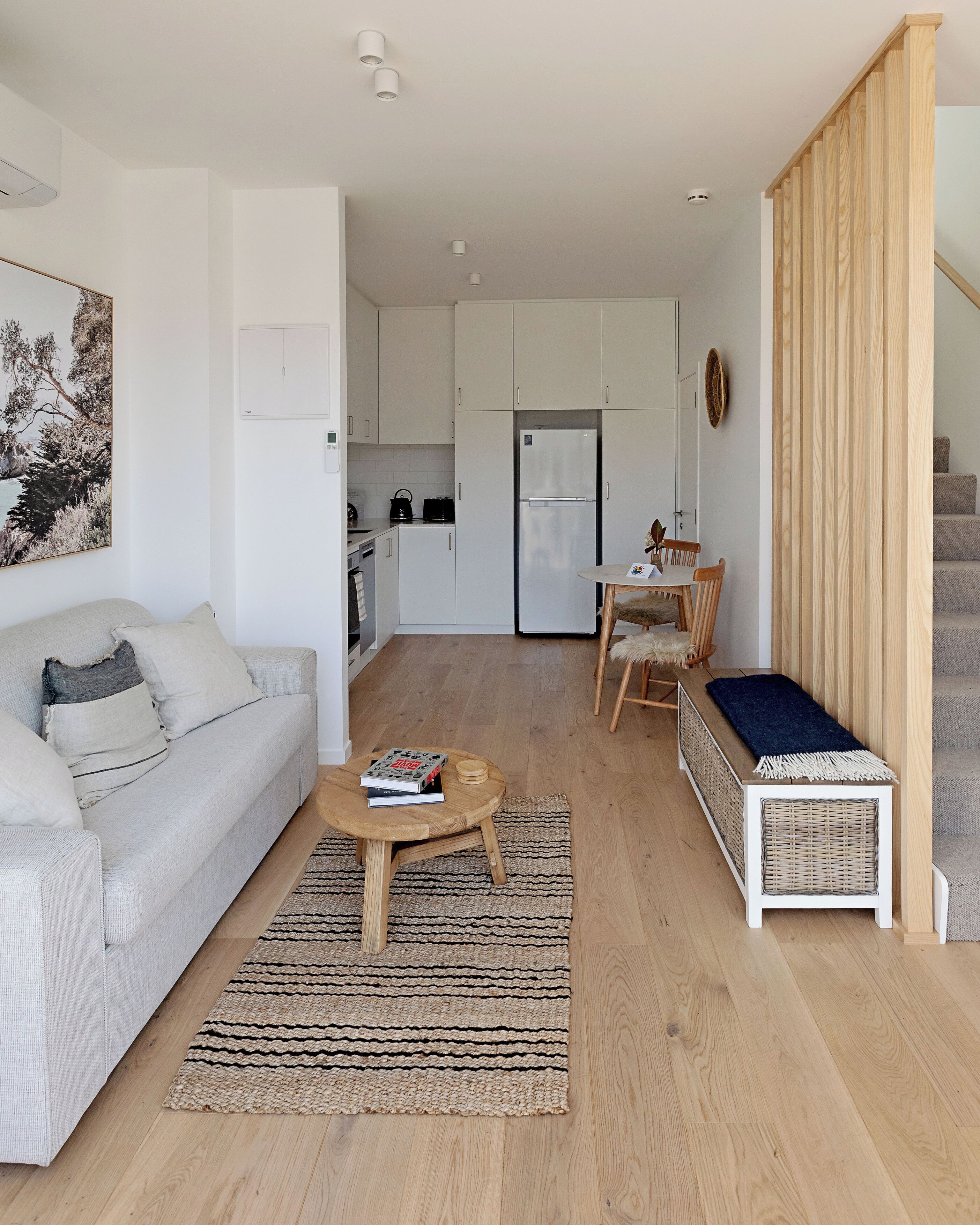 A modern living room with a sofa, a coffee table, and a dining area with a chair and table. A kitchen with white cabinets and a refrigerator is visible in the background.
