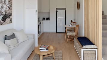 A modern living room with a sofa, a coffee table, and a dining area with a chair and table. A kitchen with white cabinets and a refrigerator is visible in the background.