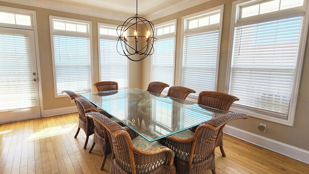 A dining room with a glass-top table surrounded by wicker chairs, large windows with blinds, and a modern pendant light. - 5 bedroom house with pool, close to center of town (Ocean Isle Beach)