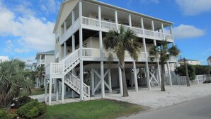A two-story beachfront building with a balcony on the upper floor, a staircase leading to the upper level, and palm trees in the foreground. - 5 bedroom house with pool, close to center of town (Ocean Isle Beach)