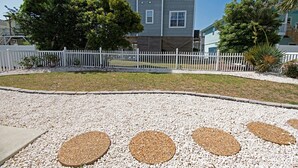 Un barrio residencial con una casa de dos pisos, cerca viva blanca y un camino de grava con piedras redondas.