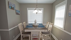 Comedor con mesa de madera, seis sillas, una araña y dos ventanas con persianas.