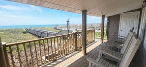 Een houten terras met een hekwerk dat uitkijkt over een strand, een houten bankje en een deur.