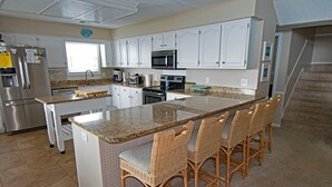 Eine Küche mit weißen Schränken, einer Granit Arbeitsplatte, einem Edelstahlkühlschrank, einer Mikrowelle und einem Toaster. An der Kochinsel stehen Korbstühle.