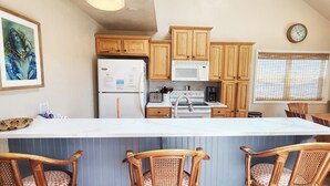 Een keuken met houten kastjes, een witte koelkast, een magnetron, een koffiezetapparaat en een eethoek met rieten stoelen.