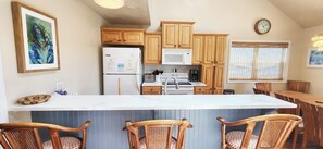 A kitchen with wooden cabinets, a white refrigerator, a microwave, a coffee maker, and a dining area with wicker chairs. - Isle Be Seeing You 4 BR Dog Friendly Canal House (Ocean Isle Beach)