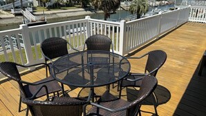 Een houten terras met een ronde tafel en vier stoelen, met uitzicht op een gracht met boten en gebouwen op de achtergrond.