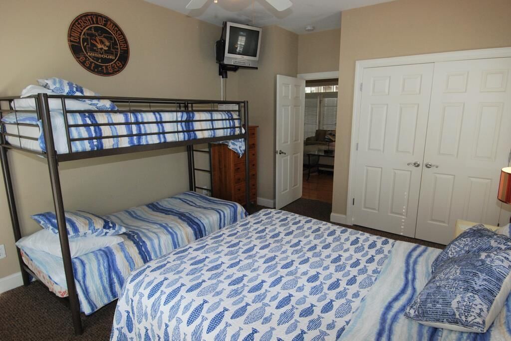 A dormitory room with bunk beds, a television, a door leading to another room, and a wall-mounted emblem.