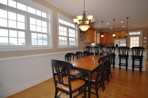 A spacious dining area with a large wooden table and black chairs, surrounded by large windows and a chandelier. - Shore Beats Work 5 BR/5BA Private Pool, Closet to Beach (Ocean Isle Beach)