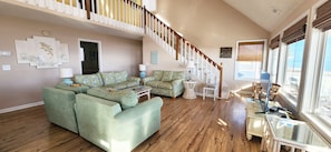 A spacious living room with a large sectional sofa, a glass coffee table, and a staircase leading to an upper level. - Stones Throw 4 BR Ocean View Beach House (Ocean Isle Beach)