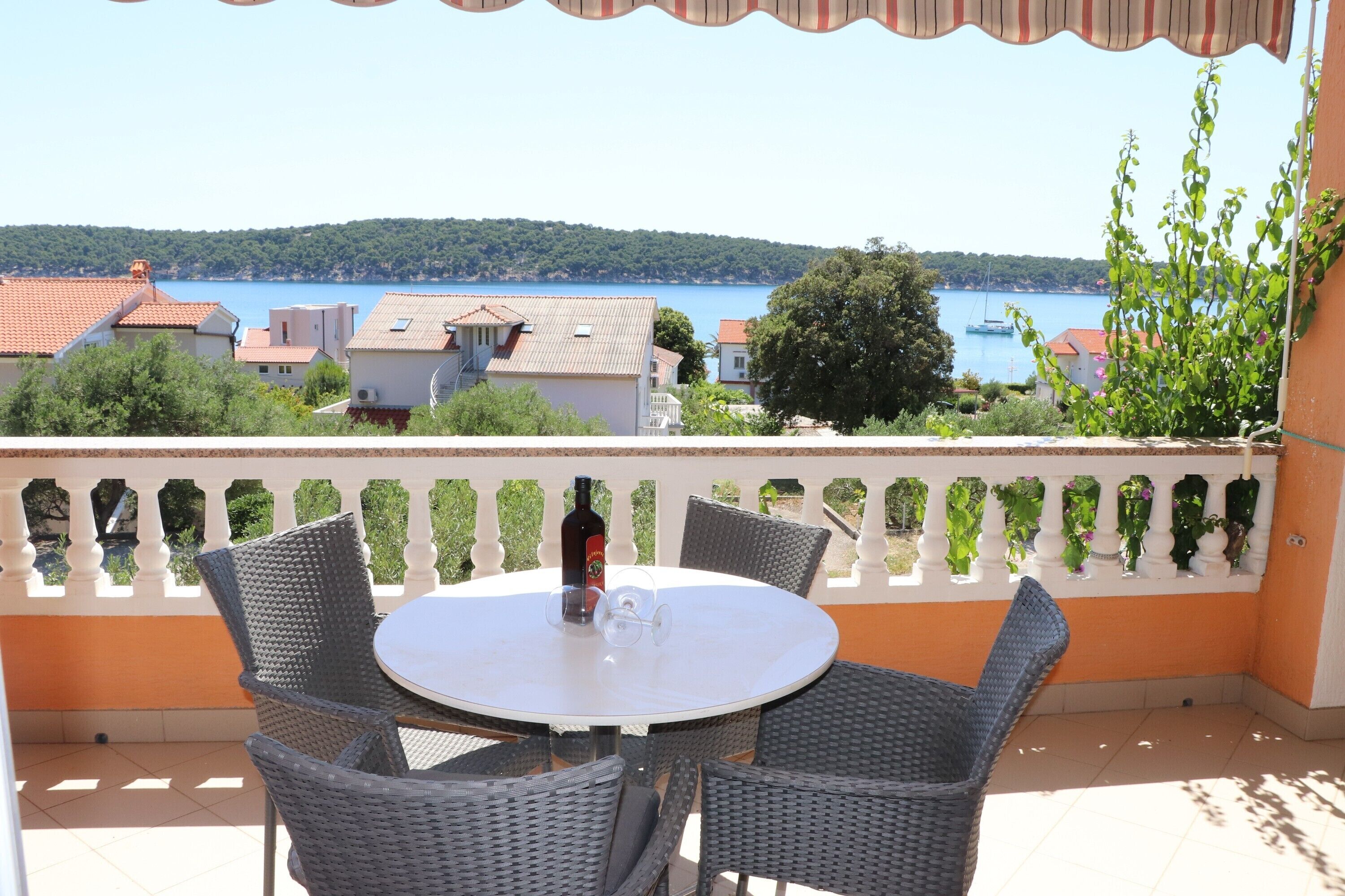 Una mesa redonda con sillas de mimbre en un balcón con vistas a un lago donde se ven barcos y casas al fondo.