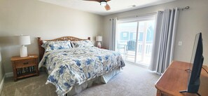 A bedroom with a bed, a nightstand, a TV stand, and a large window with curtains. - A Southern Escape 5 BR Summer Place Home (Ocean Isle Beach)