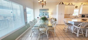A dining area with a glass table, white chairs, and a chandelier. - Oceanfront 6 bedroom beach house (Ocean Isle Beach)