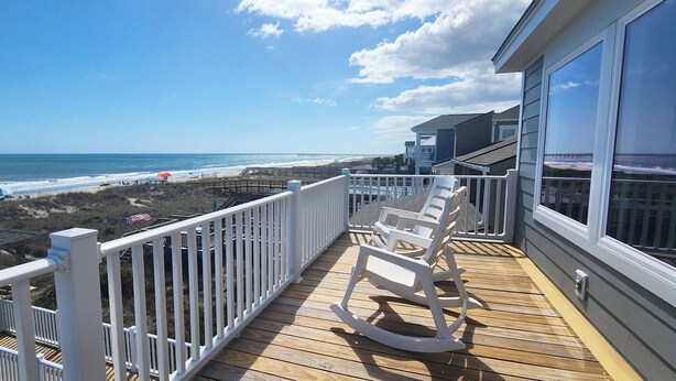 Un balcon avec vue sur la plage et l’océan, des fauteuils à bascule blancs et une terrasse en bois.