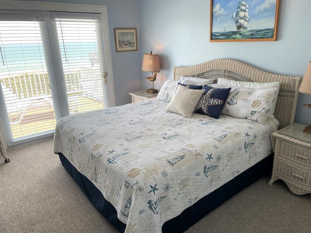 A bedroom with a bed, a nightstand, a lamp, a painting, and a view of the ocean through a door.