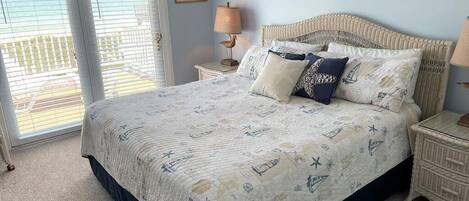 A bedroom with a bed, a nightstand, a lamp, a painting, and a view of the ocean through a door.
