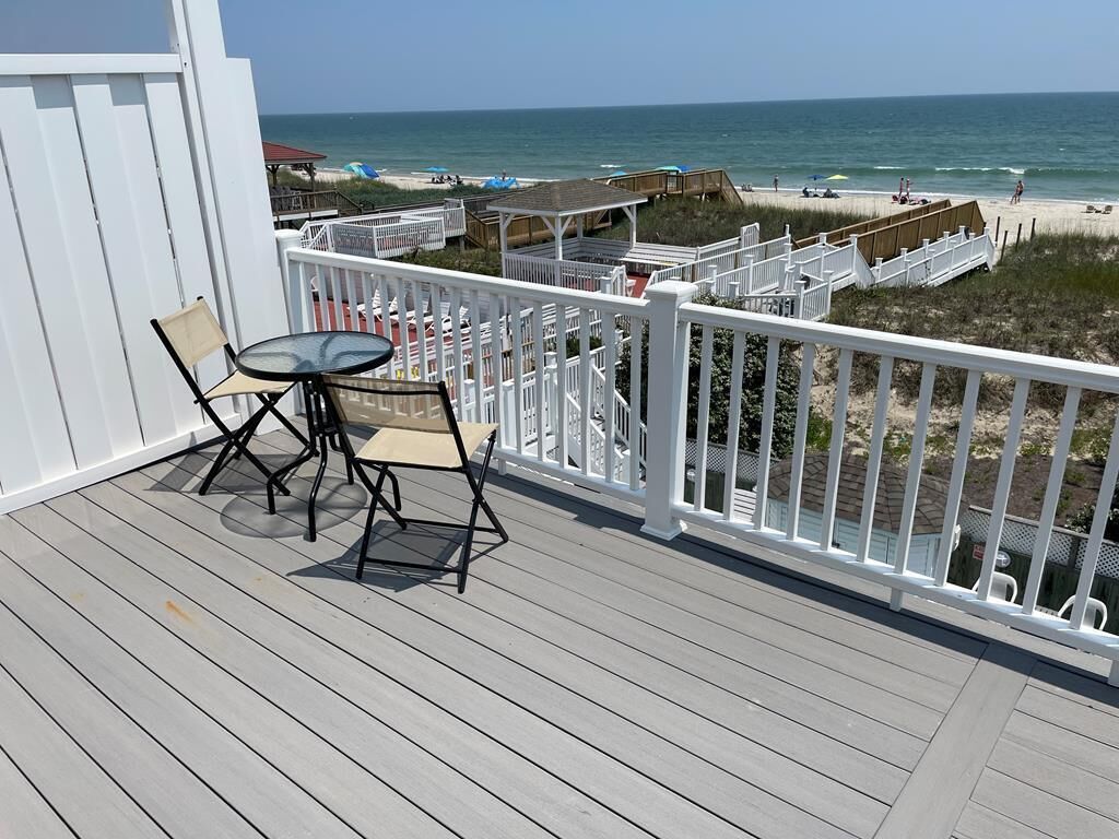 A wooden deck with a table and chairs, overlooking a beach with umbrellas and a wooden walkway.