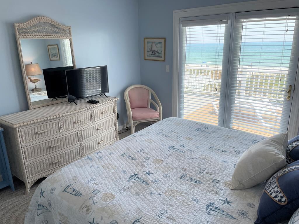 A bedroom with a bed, dresser, TV, chair, and a view of the ocean through the window.