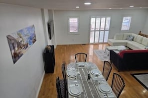 A dining area with a long table set for six, surrounded by chairs, in a room with a white sofa and a red blanket on a couch.