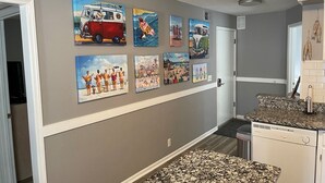Un pasillo decorado con varias obras de arte con temática de playa, una cocina equipada con lavavajillas y una puerta que da a otra habitación.