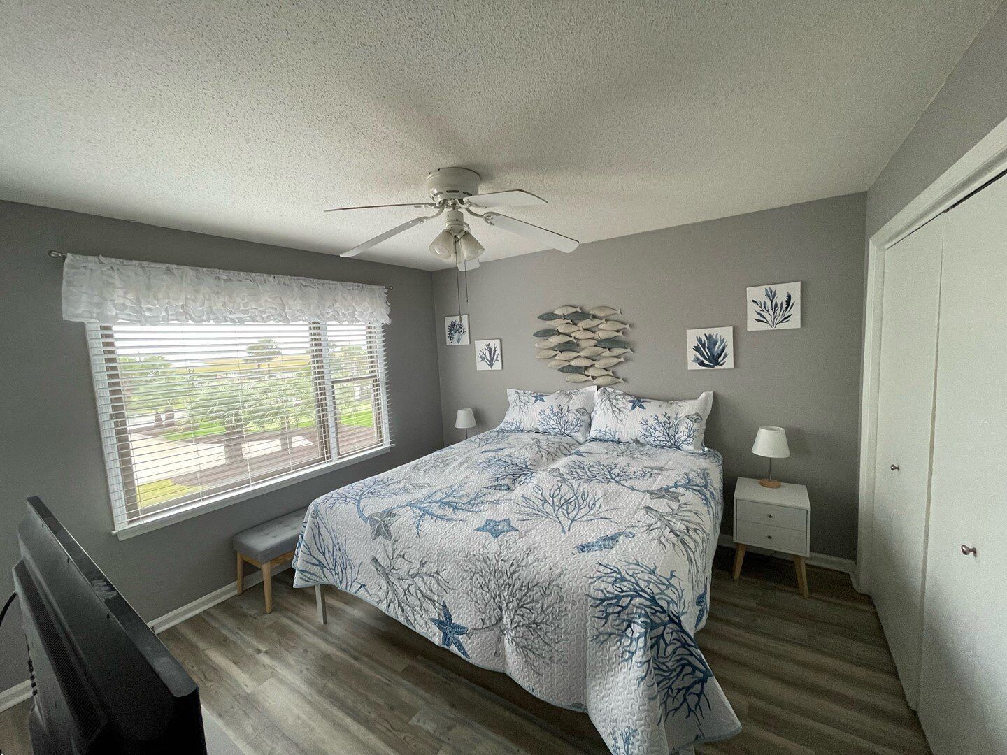 A bedroom with a bed, a ceiling fan, a window with blinds, and a wall with framed pictures.