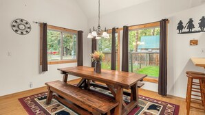 A dining area with a wooden table and benches, large windows with curtains, and a hanging light fixture. - Cozy cabin, dogs ok, hot tub, & fireplace (Leavenworth)