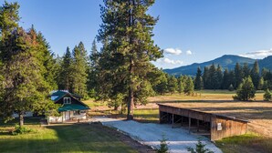 A rustic cabin with a green roof, a covered porch, and a gravel driveway leading to a wooden dock. - A cozy dog friendly cabin, sleeps 6 with a hot tub (Leavenworth)