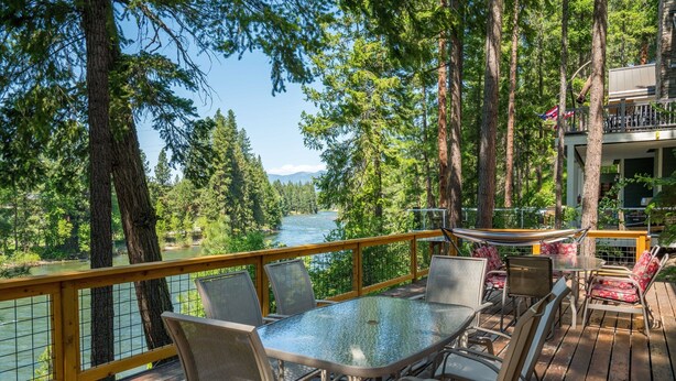 Una terrazza con tavolo e sedie in vetro affacciata su un fiume, circondata da alberi e una ringhiera in legno.