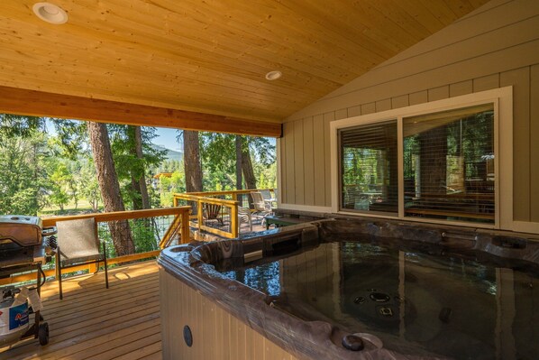 Una terraza de madera con un jacuzzi, mobiliario exterior y vistas a árboles.