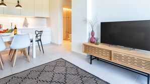 Ein modernes Wohnzimmer mit großem Flachbildfernseher auf einer Holzkonsole, einem Essbereich mit weißem Tisch und Stühlen sowie einer Küche mit weißen Schränken und Pendelleuchten.