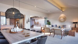 Ein modernes Interieur mit Essbereich, der einen langen Tisch und Stühle umfasst, einem Wohnzimmer mit Sofa und Couchtisch sowie einer Küche im Hintergrund.