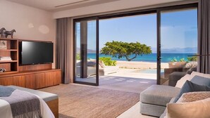 Ein modernes Wohnzimmer mit einem großen Flachbildfernseher, einer Couch und einem Couchtisch. Eine Glastür öffnet sich zu einem Balkon mit Blick auf das Meer und einen Baum.