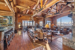 A log cabin interior with a wooden bar, seating area, and large windows offering mountain views.