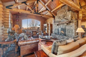 A log cabin interior with a stone fireplace, leather sofas, and a large window overlooking the landscape.