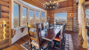 Interior de una cabaña de madera con una mesa de comedor, sillas, una araña y grandes ventanas con vistas a un paisaje montañoso.