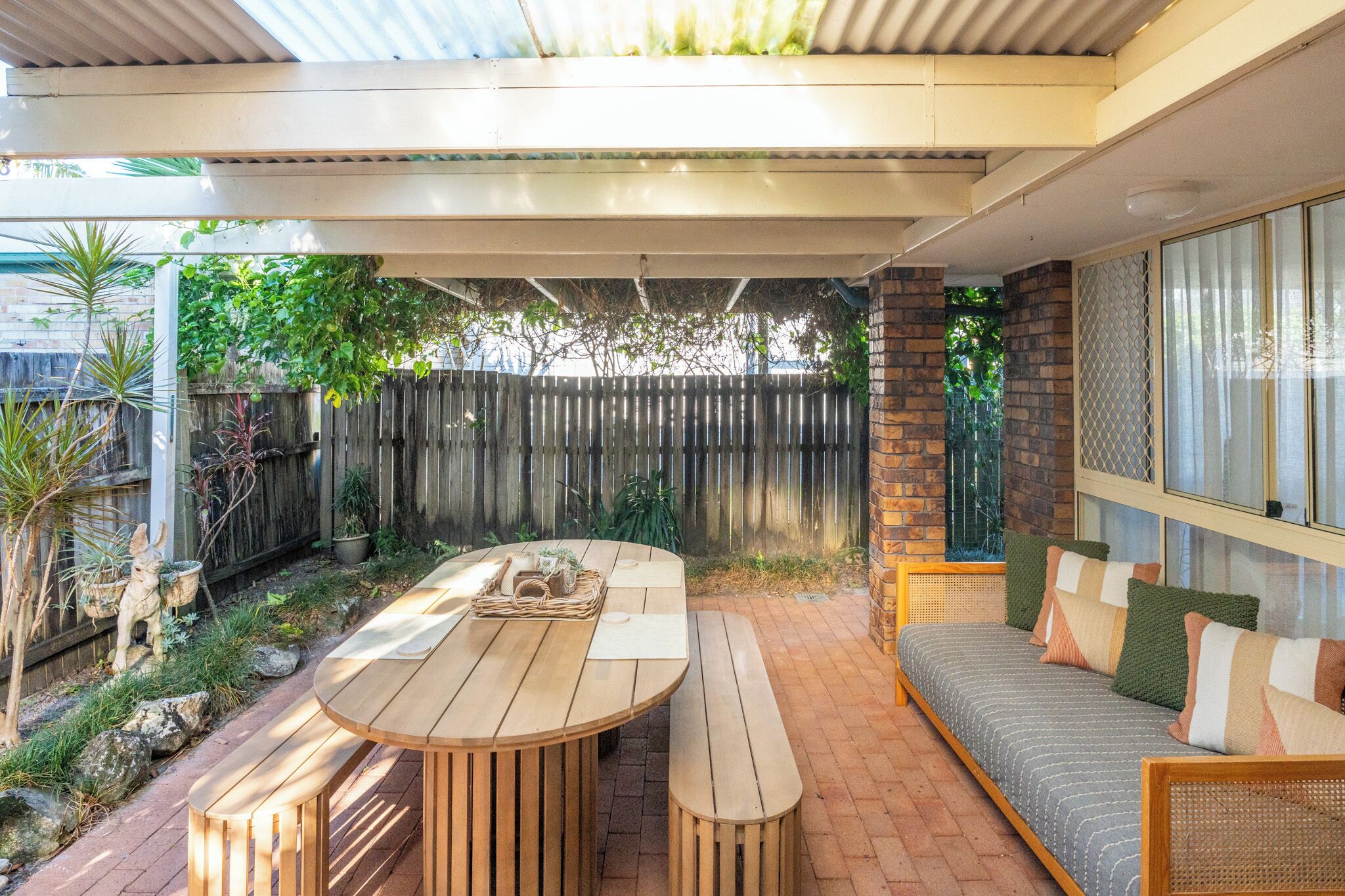 A covered patio with a wooden table and benches, a cushioned seating area, and a view of a wooden fence and greenery.
