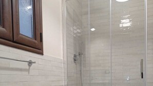 Un bagno con doccia vetrata, pareti piastrellate di bianco, un armadietto in legno e un piano di lavoro in marmo.