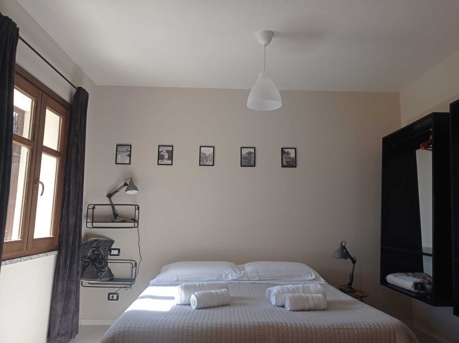 Un dormitorio con cama, lámpara colgante, estante en la pared y ventana con cortinas.