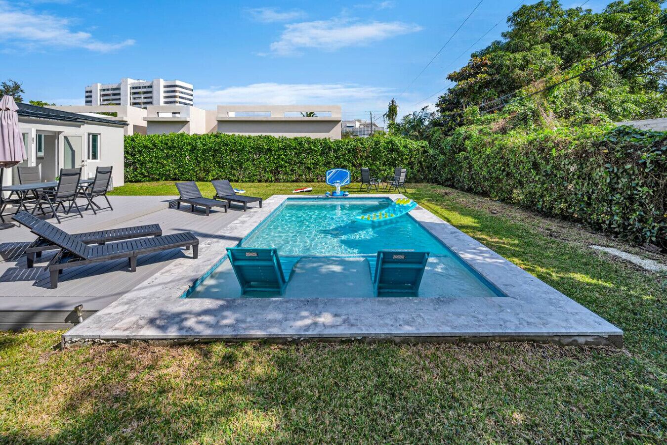 A rectangular swimming pool with a diving board and lounge chairs, surrounded by a grassy area and a hedge.