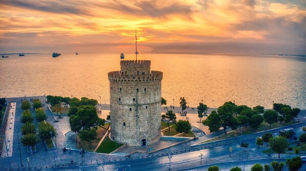 Una torre histórica con base circular y techo cónico, rodeada de una zona pavimentada con árboles y vistas al agua.