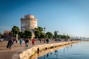 Una torre histórica de base circular y parte superior cónica, rodeada por un paseo marítimo con árboles y farolas.