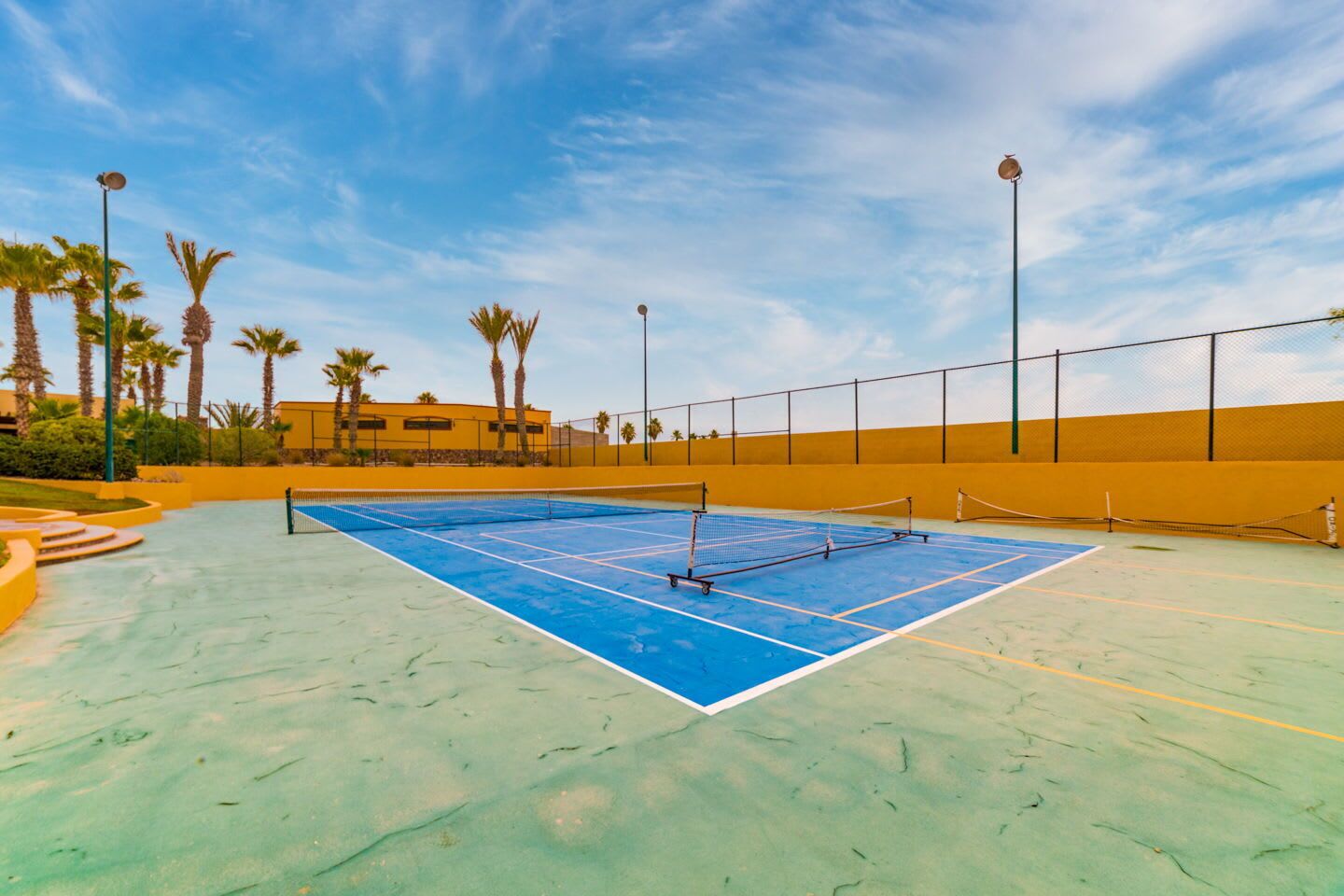 Una cancha de tenis al aire libre con red, rodeada de palmeras y un edificio amarillo al fondo.