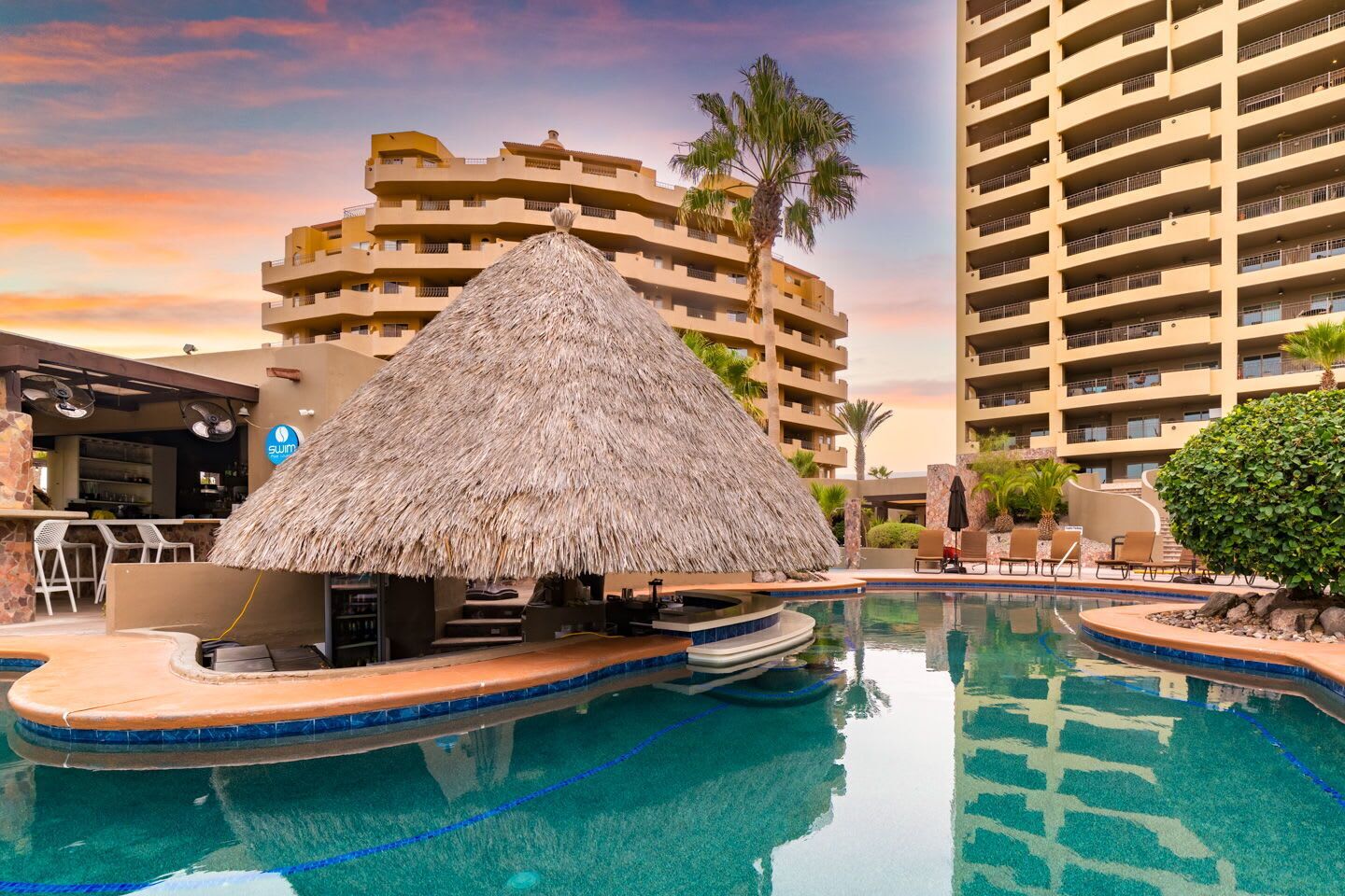 A swimming pool with a thatched-roof bar area, surrounded by palm trees and a high-rise building in the background.