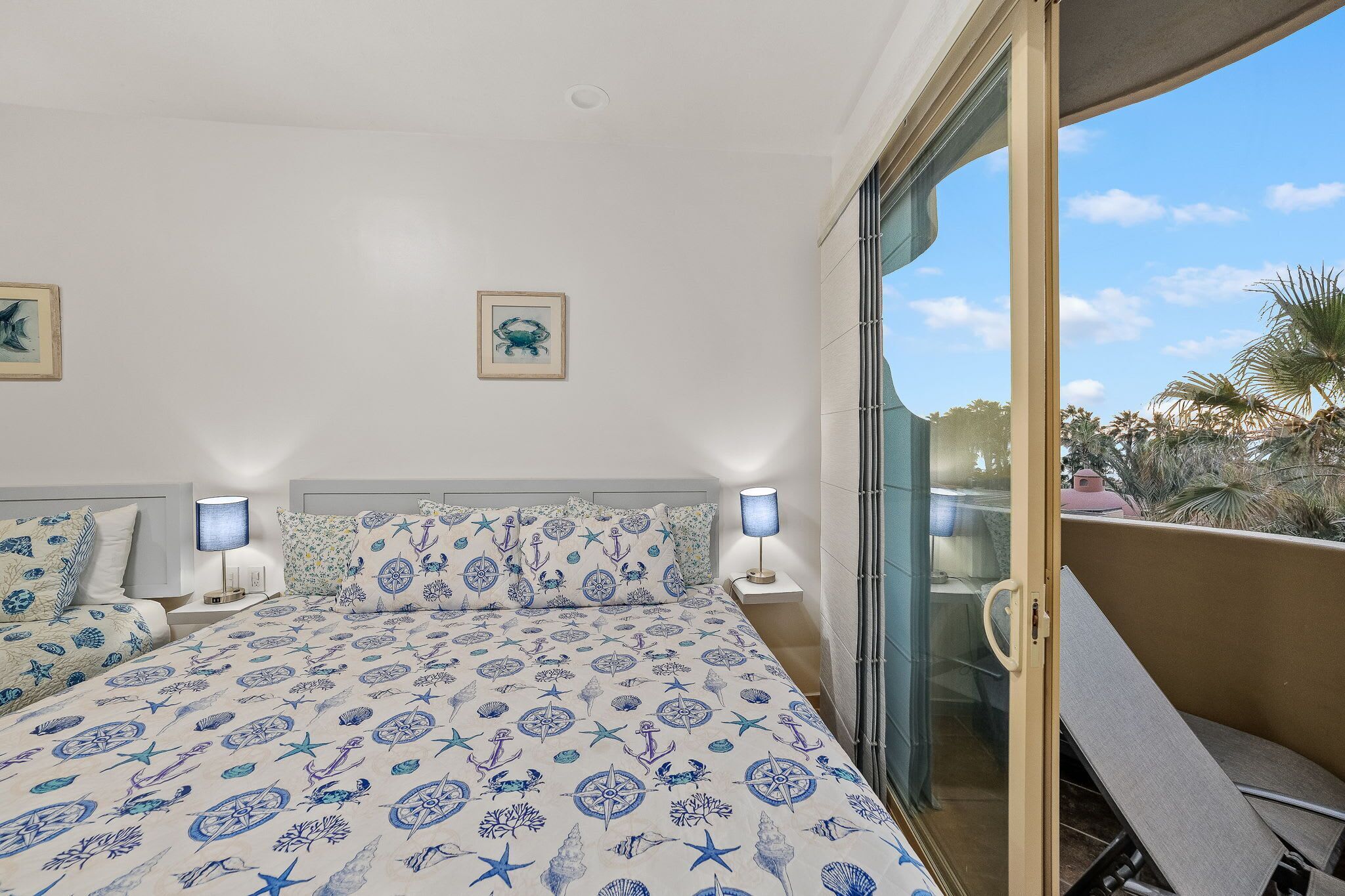 Una camera da letto con un letto e una coperta a tema nautico, due abat-jour e vista su palme attraverso una porta scorrevole in vetro.