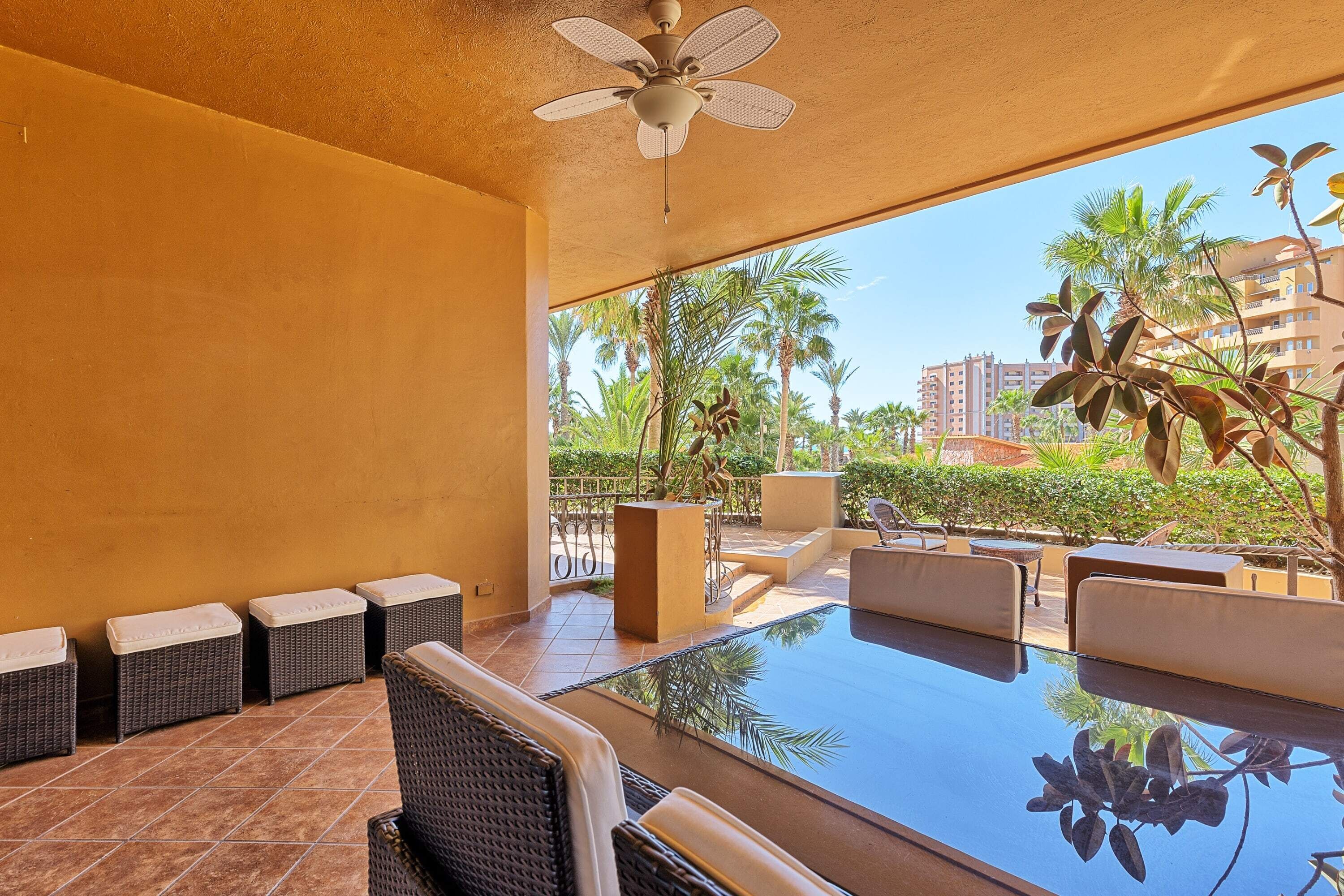 A poolside area with wicker furniture, a ceiling fan, and a view of palm trees and buildings.