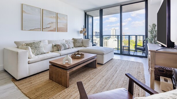 A modern living room with a white sofa, a wooden coffee table, and a large window offering a city view. - High Rise Condo Water Views Walker's Paradise (Miami)