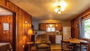 Un accogliente interno di una baita con pareti in legno, una zona pranzo con tavolo e sedie, un divano, una lampada, e una cucina con fornello, microonde e frigorifero.