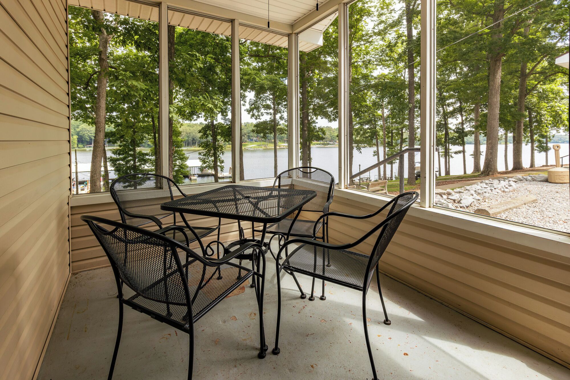 Un porche cubierto con una mesa y sillas, amplias ventanas que ofrecen vista a un lago y árboles.