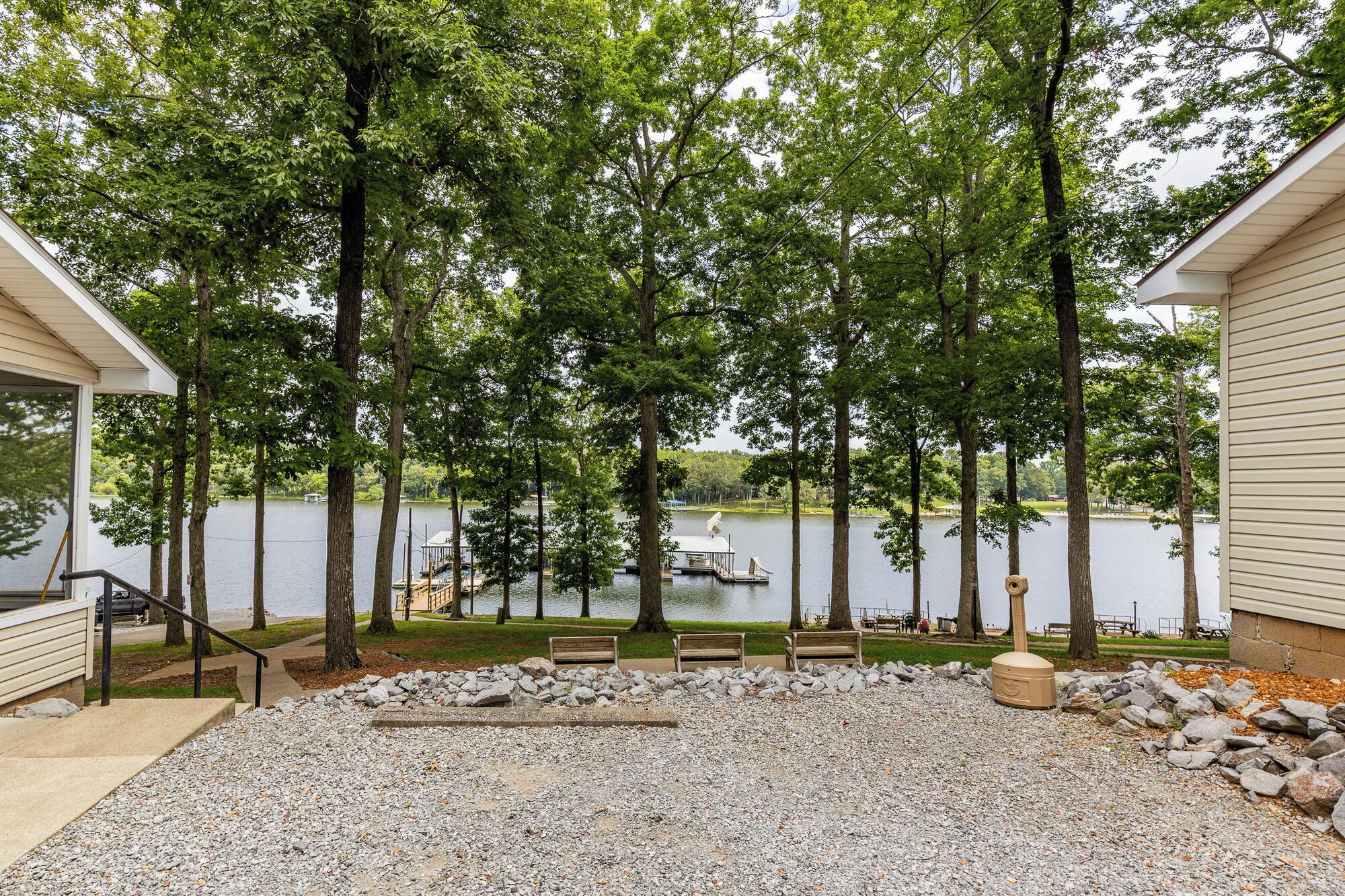 A lakeside property with a gravel patio, wooden benches, and a dock with a boat.